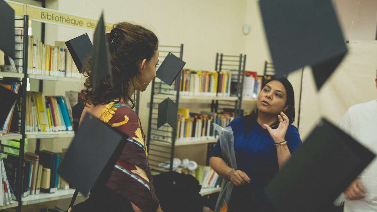 Conversación entre dos participantes del EXPYLAB en la biblioteca de la Alianza Francesa durante una sesión de trabajo, con paneles geométricos en primer plano