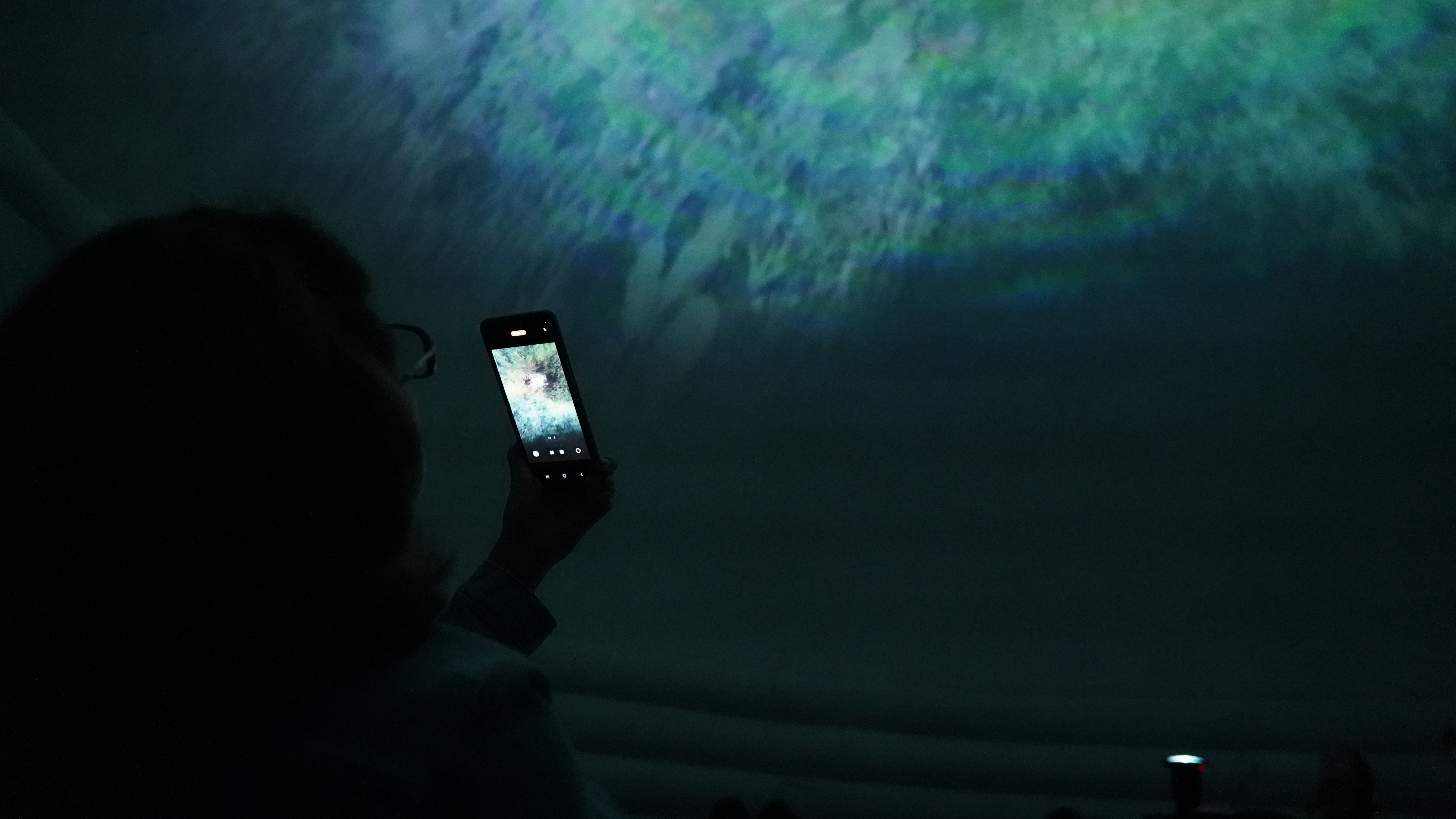 Participante de EXPYLAB documenta con su celular una proyección inmersiva sobre cúpula/planetario en tonos azules y verdes