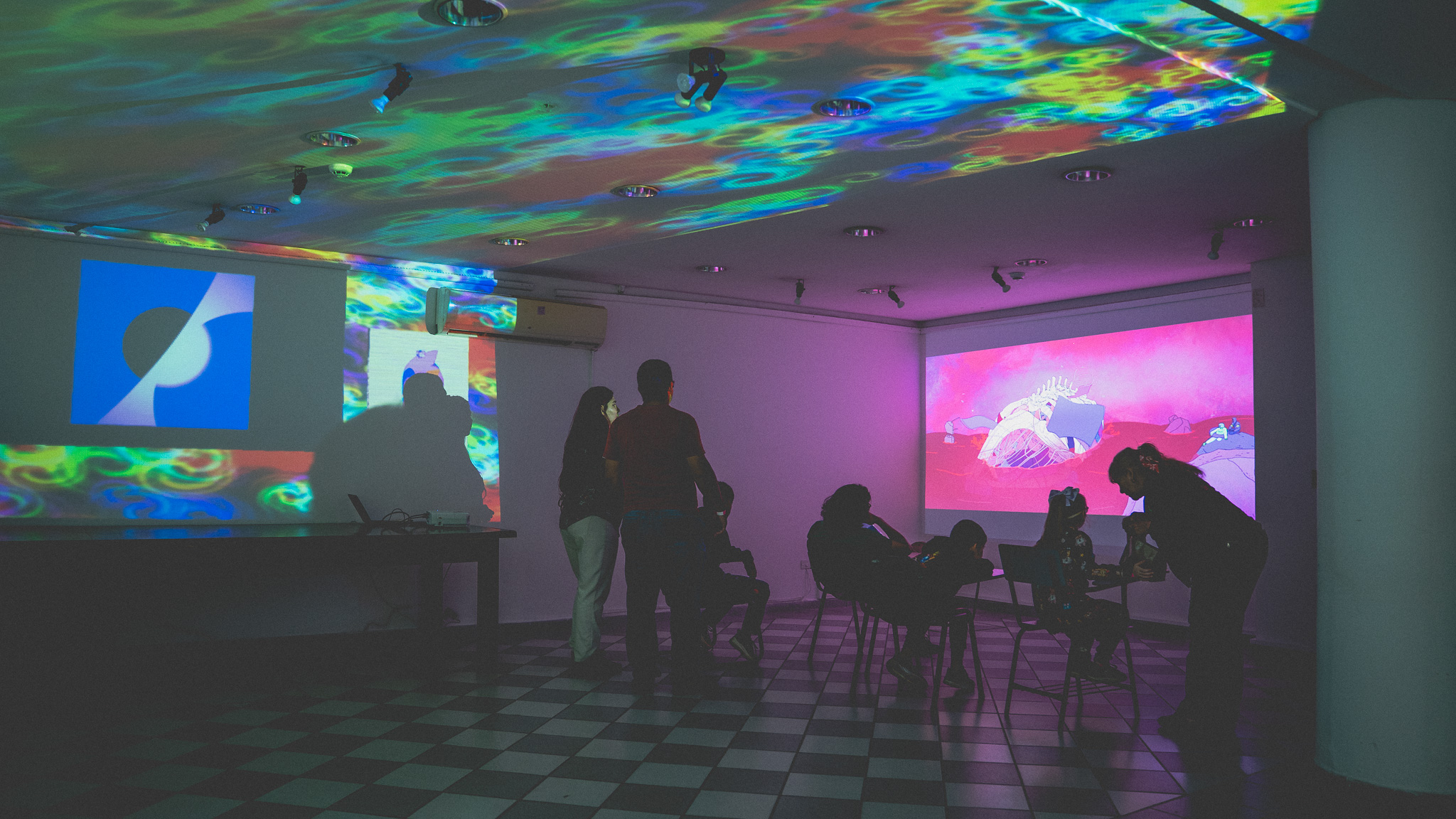 Sala inmersiva del EXPYLAB con proyecciones envolventes — patrones psicodélicos en el cielorraso y dos pantallas paralelas con animaciones, participantes silueteados sobre piso ajedrezado
