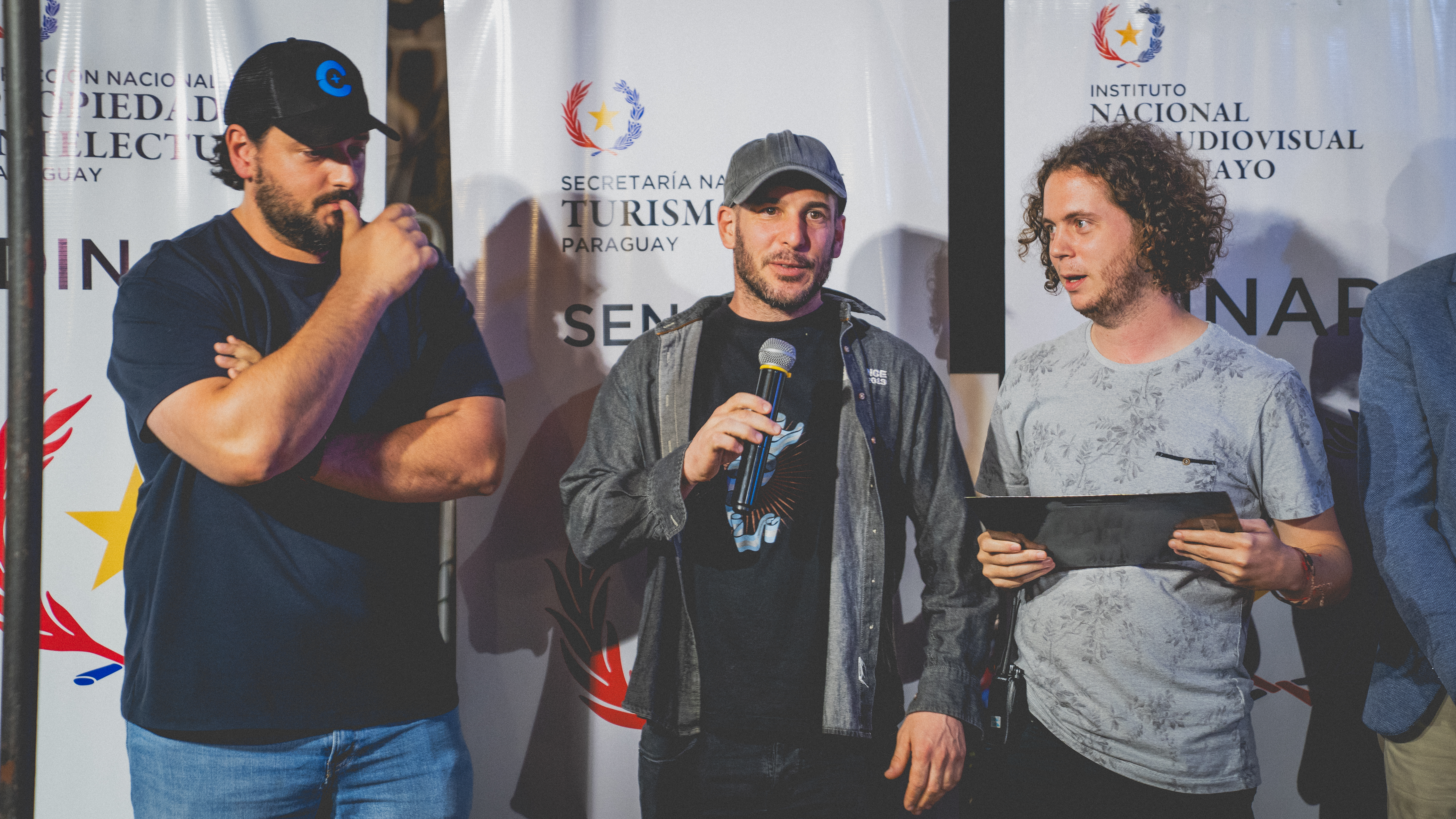 Tres participantes del EXPYLAB en escenario durante la presentación de proyectos — uno habla por micrófono, banners de auspiciantes (Senatur, INAP, Propiedad Intelectual) al fondo